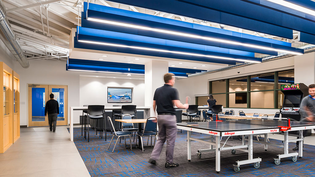 People playing ping pong in a room with tables and chairs.