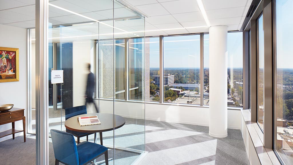 A room with a glass wall and a table with chairs and a person standing in the distance.