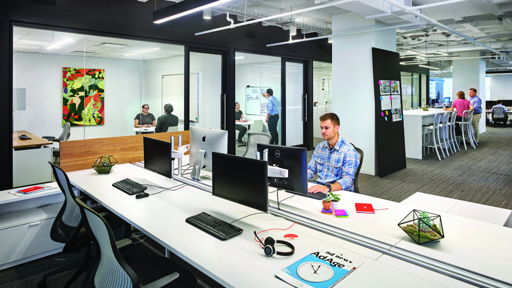 A person sitting at a table with computers and other people in the background.