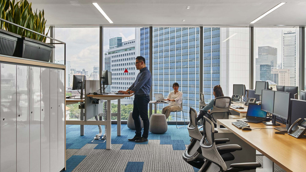 A person standing in front of a desk with a computer in front of a large window.