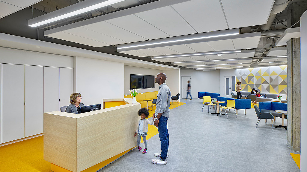 A person and a child walking in a room with tables and chairs.