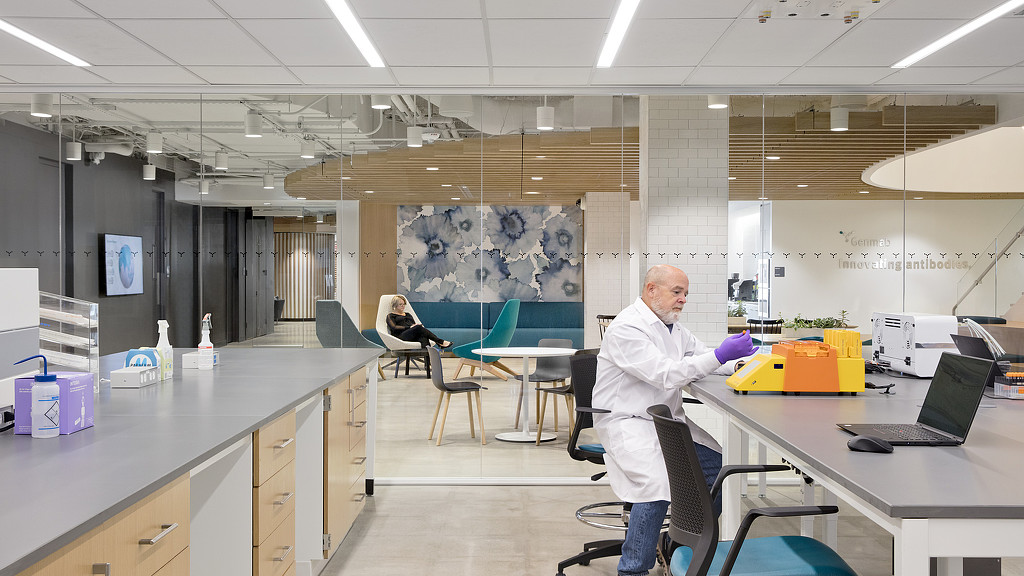 A person wearing a white coat and gloves in a room with other people.