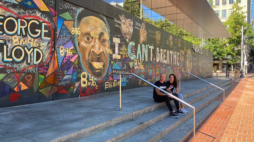 A man and woman sitting on a sidewalk next to a wall with graffiti.