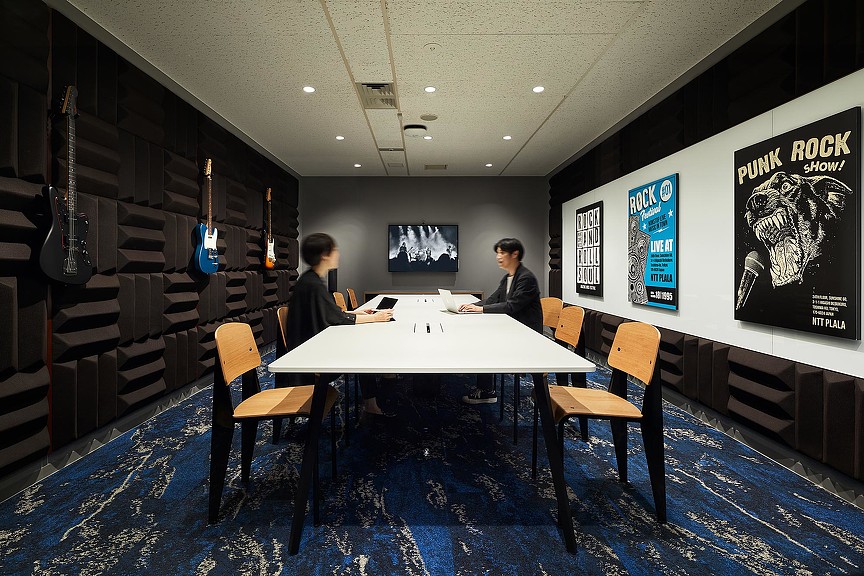 A couple of men sitting at a table in a room with a large screen and chairs.