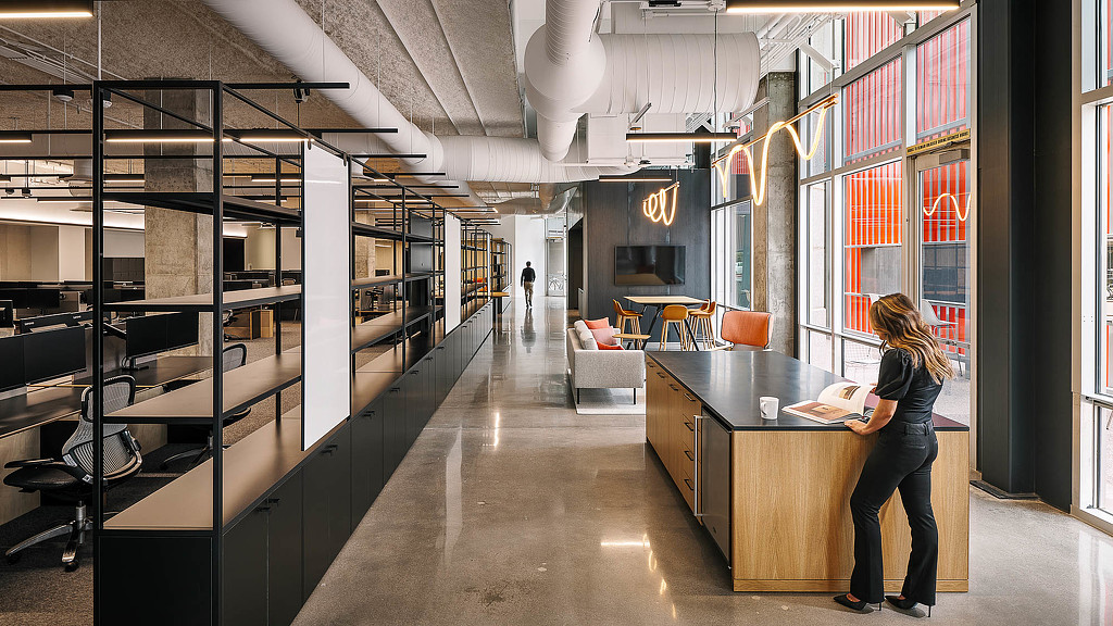 A person standing at a desk in a room with shelves and a table.
