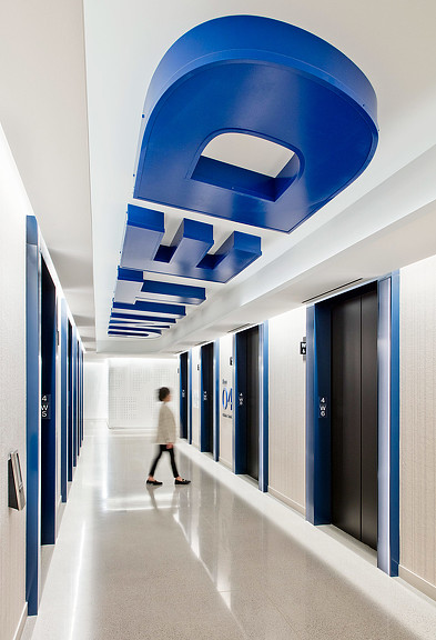 A person walking in a hallway to elevators with environmental graphic design on the ceiling at United Airlines lounge