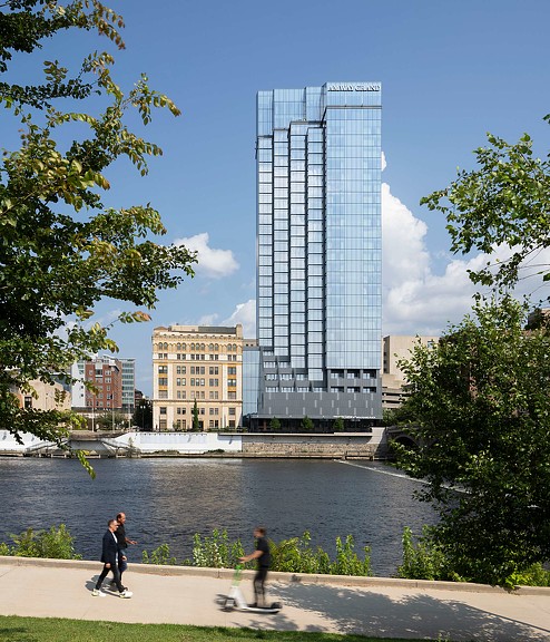 A couple of people skateboarding by a river and the tall glass facade of Amway Grand Plaza Hotel