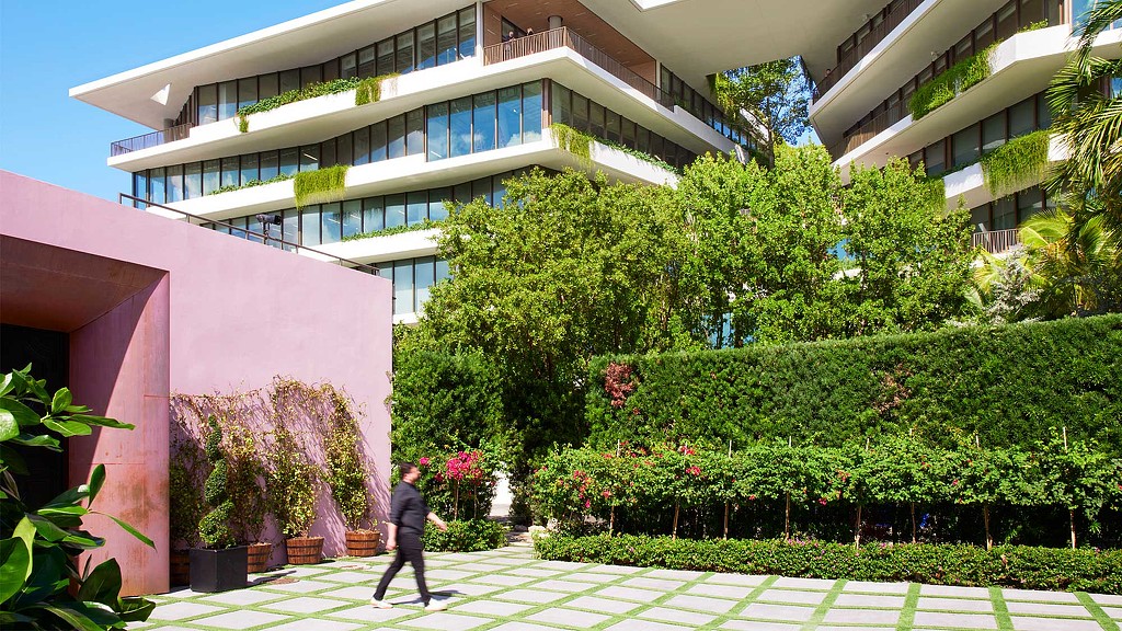 A person walking in front of a building with trees and bushes.