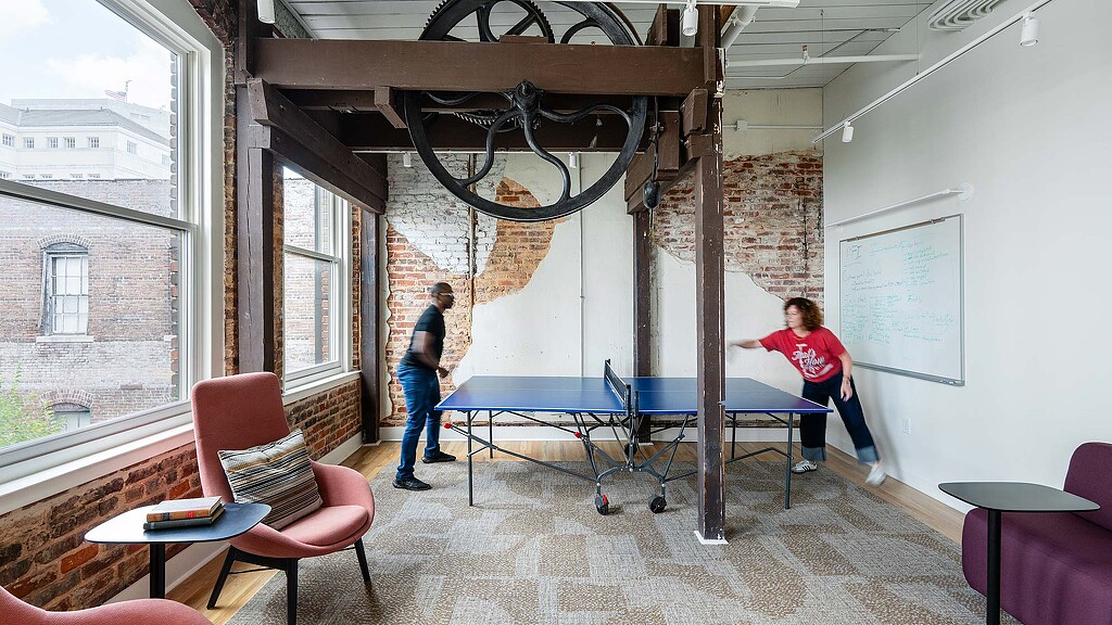 A couple of people playing ping pong in a room with a brick wall and a large window.