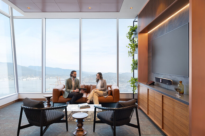 A man and woman sitting on a couch in a room with large windows.