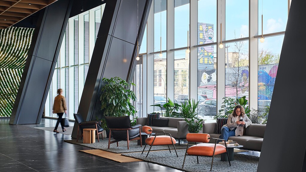 A person sitting on a couch in a room with windows and a person standing.