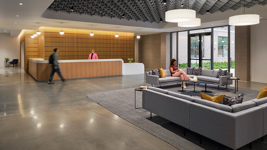 A woman sitting on a couch in a large room with a man and a woman walking by.