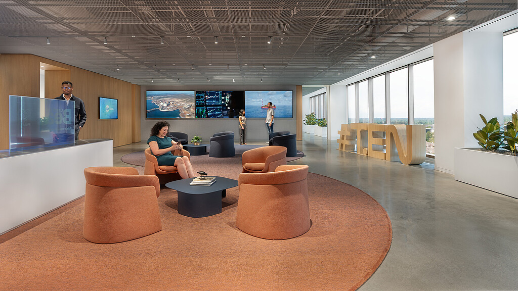 A person sitting on a couch in a room with people standing and a large screen.