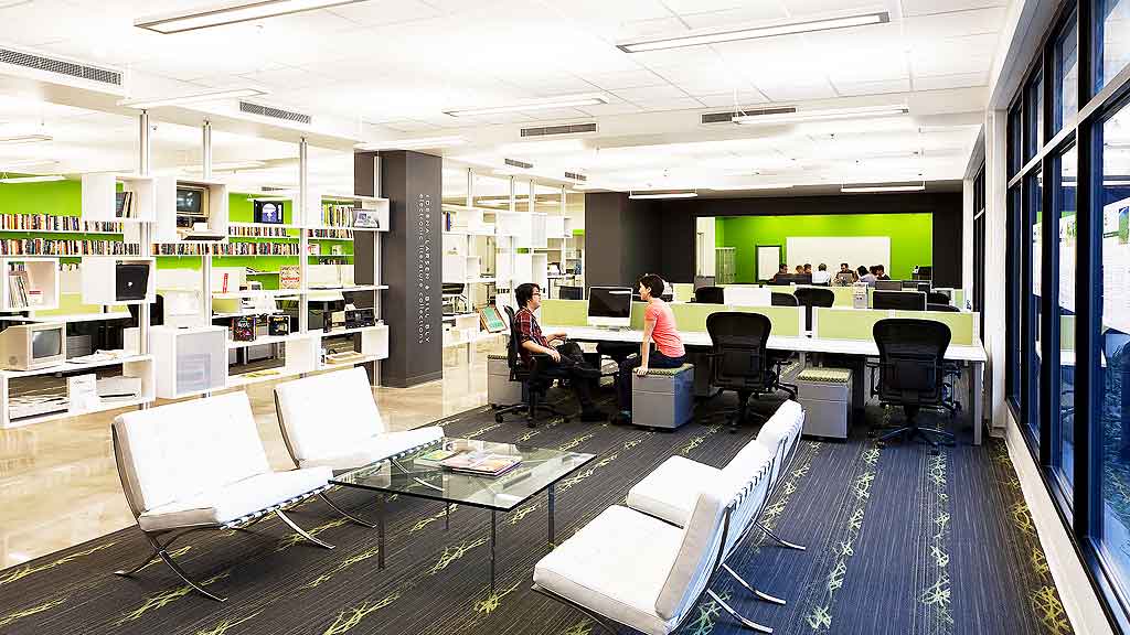 A couple of people sitting at a table in a room with a large screen and chairs.