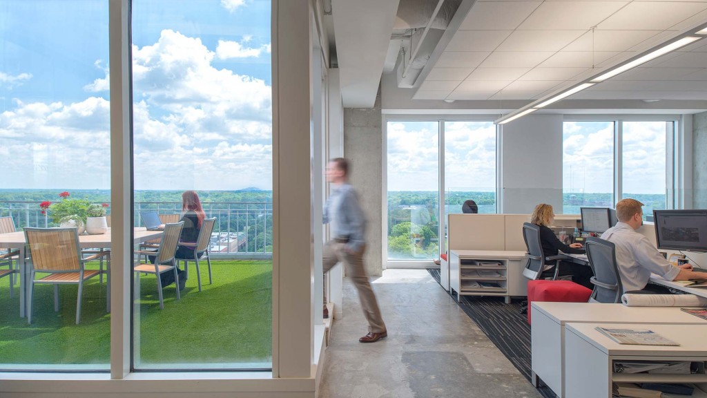 A person walking in a room with windows and a couple of people sitting at a desk with computers.