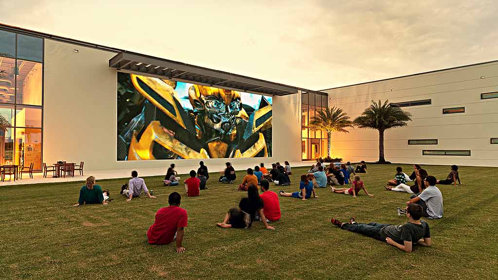 A group of people sitting on the grass in front of a large screen.