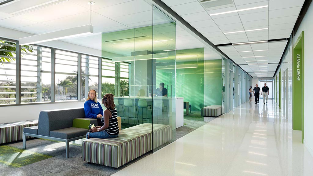A couple of women sitting on a couch in a large room with glass walls and a large window.