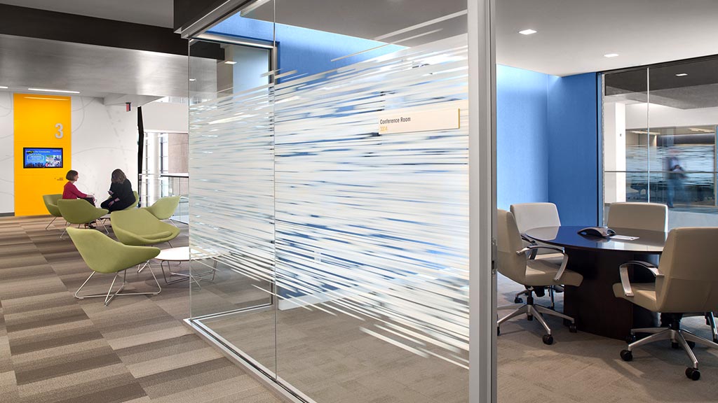 A room with a glass wall and a table with chairs and a person sitting at a desk.
