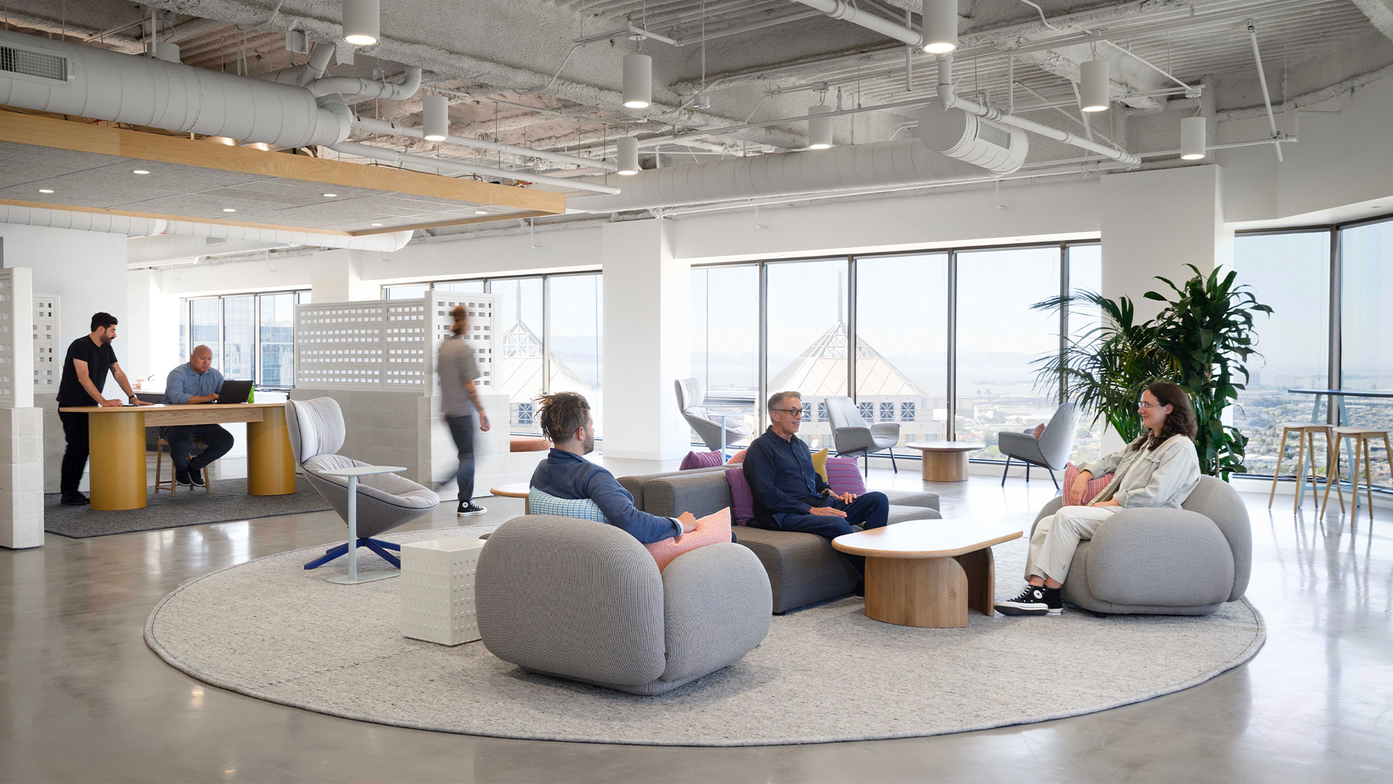 A group of people sitting in a room with windows and a person standing.