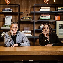 A man and a woman sitting at a desk in front of a book shelf.