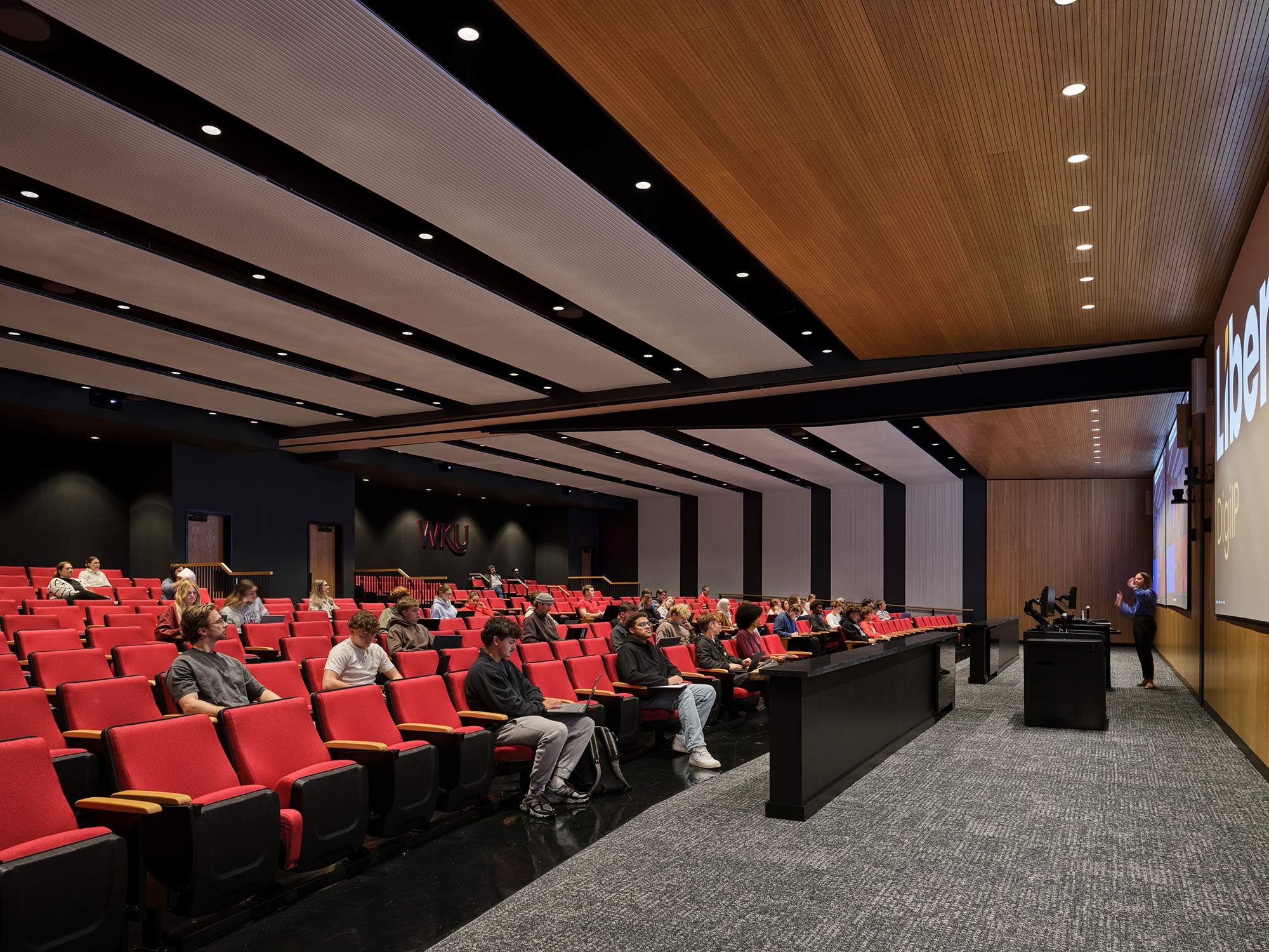 A group of people sitting in a room with a stage and a person standing in front of them.