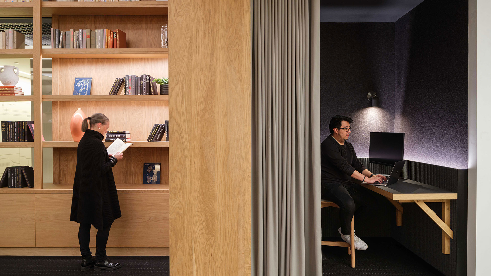 A person standing in front of a desk with a laptop and a person sitting at a computer.