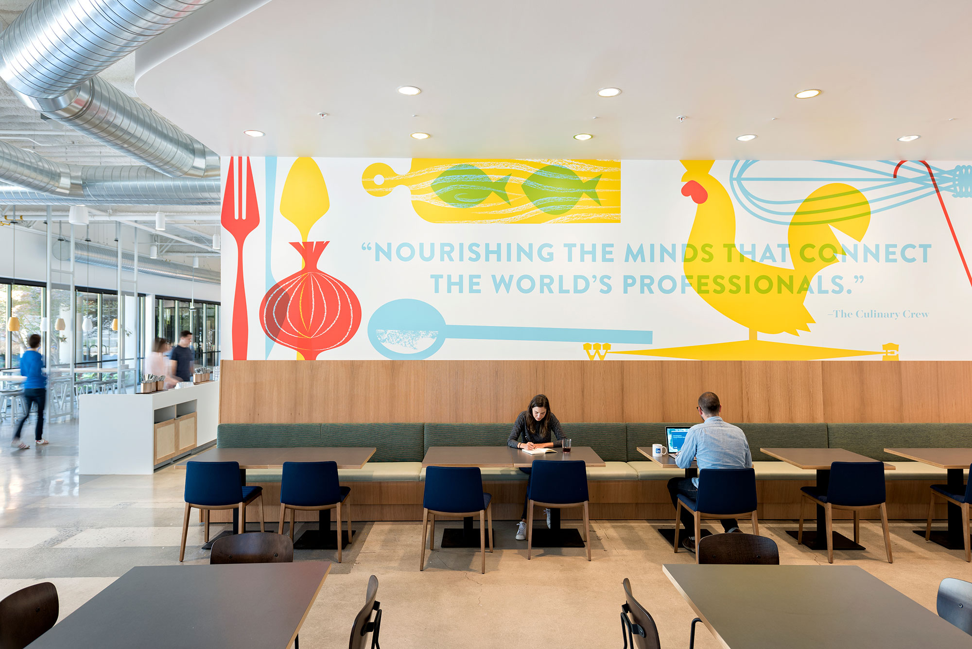 A group of people sitting at a table in a large room with a large banner.