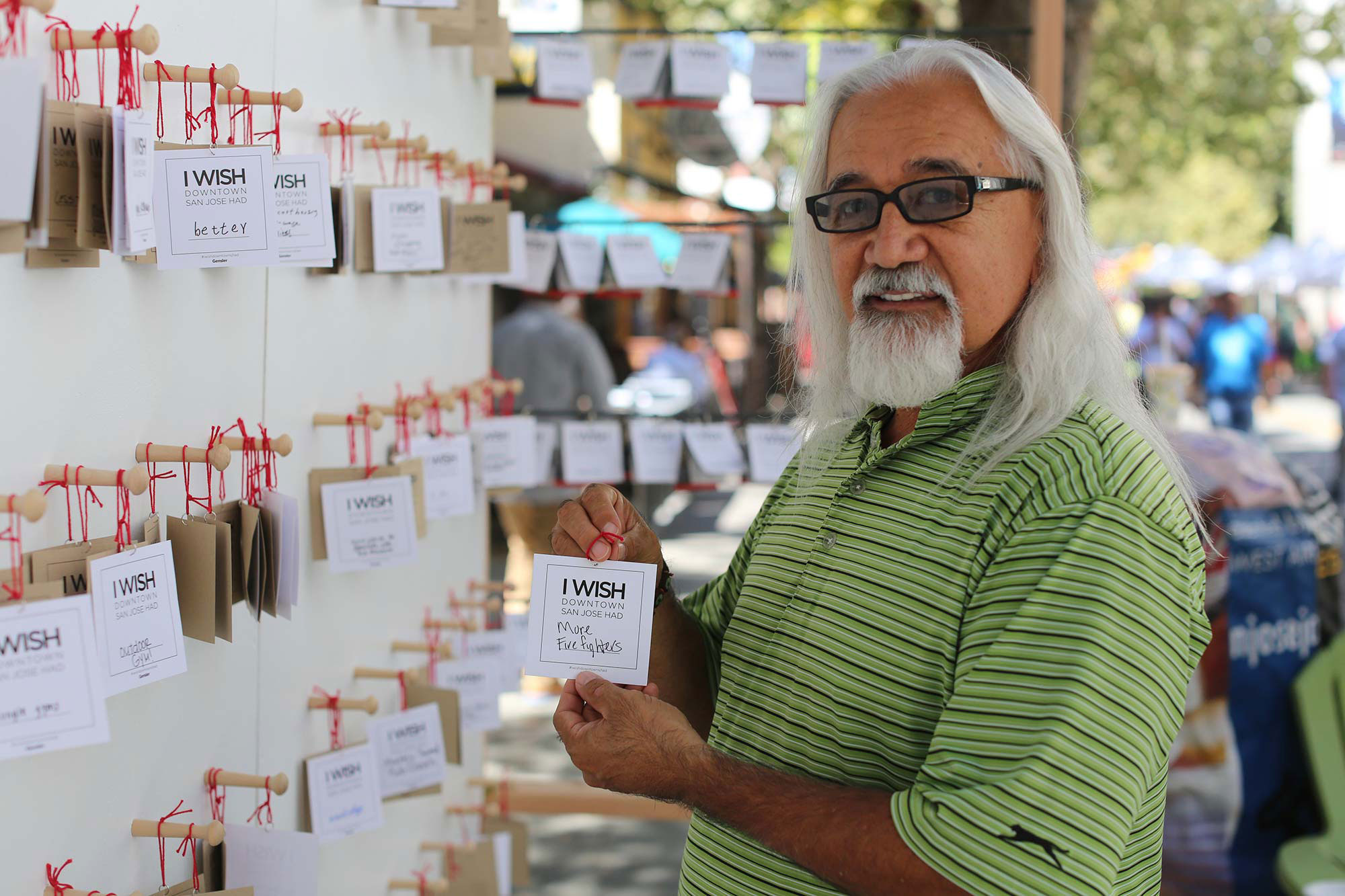 Man holding a notecard that reads "I wish San Jose had more firefighters" in front of an outdoor wall with similar notecards and wishes.