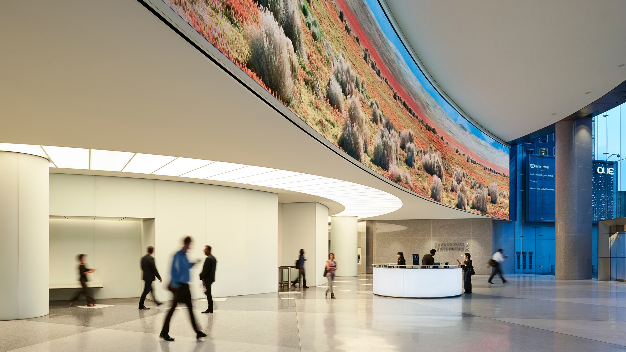 People walking through an open office tower lobby, beneath a 130-foot wide digital display showing landscape images.