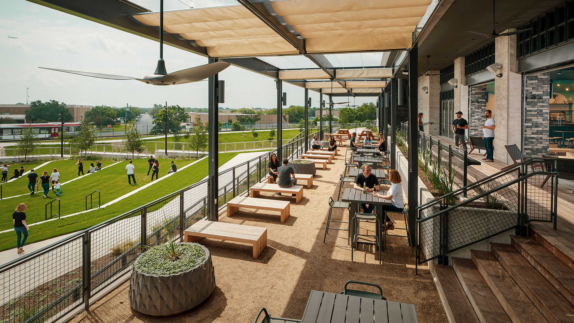 People sitting at tables in a large open area with a grassy field and people walking around.