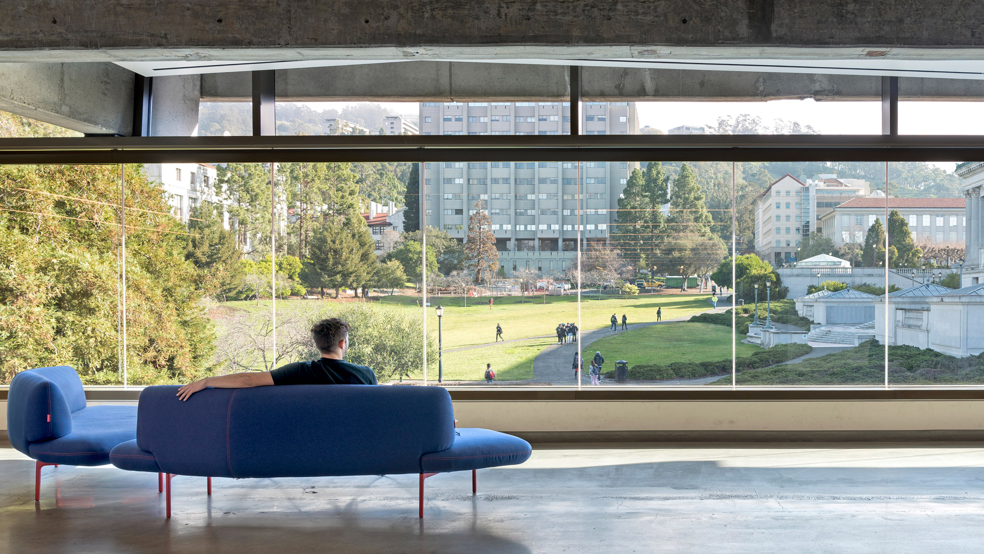 A person sits on a couch in front of a wall of floor to ceiling windows looking out on a university campus lawn and academic buildings.