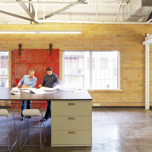 A couple of men sitting at a table in a room with a brick wall and a window.
