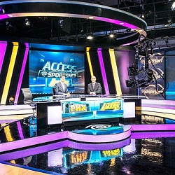 A couple of men standing behind a counter with a large screen behind them.