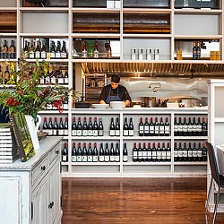 A person behind a counter with bottles of wine.