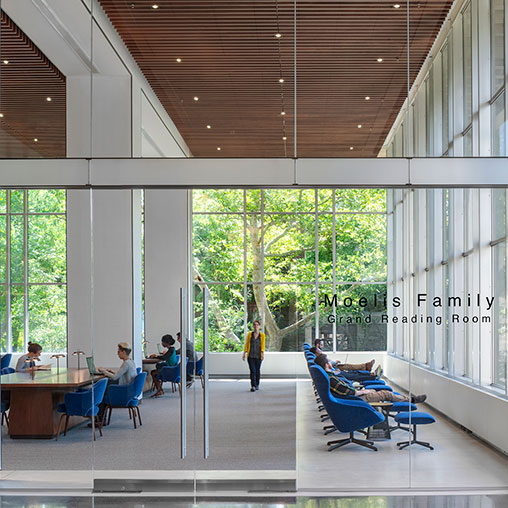 A group of people sitting in a room with windows and a person walking.