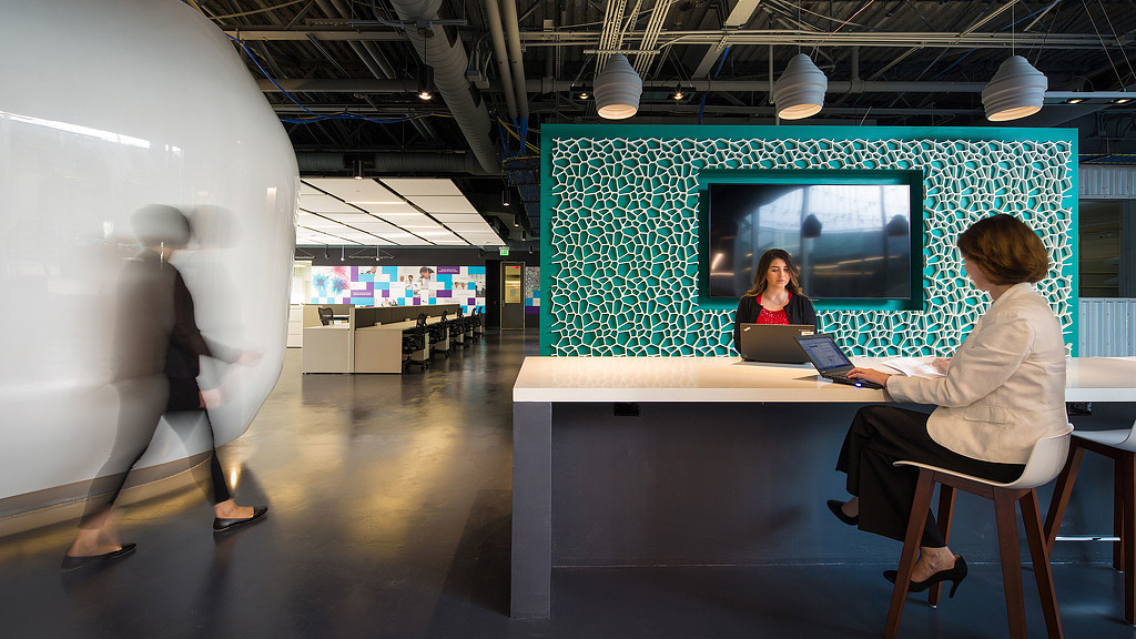 A man and woman working on a laptop in a room with a large screen.