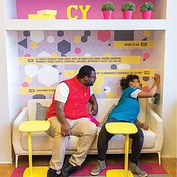 A man and a woman sitting on yellow chairs in front of a wall with colorful tiles.