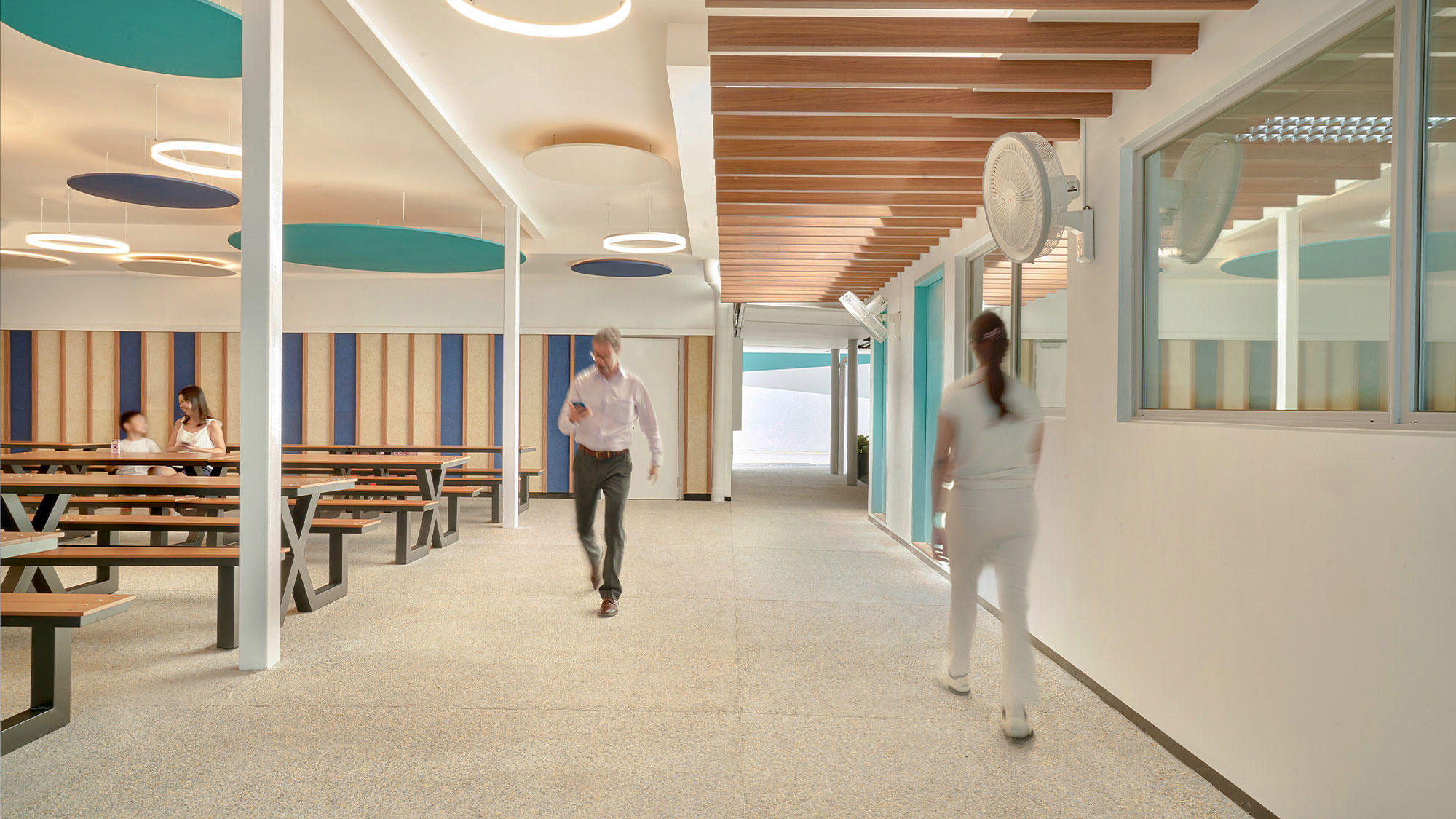 A man and a woman walking in a room with a fan and a table.
