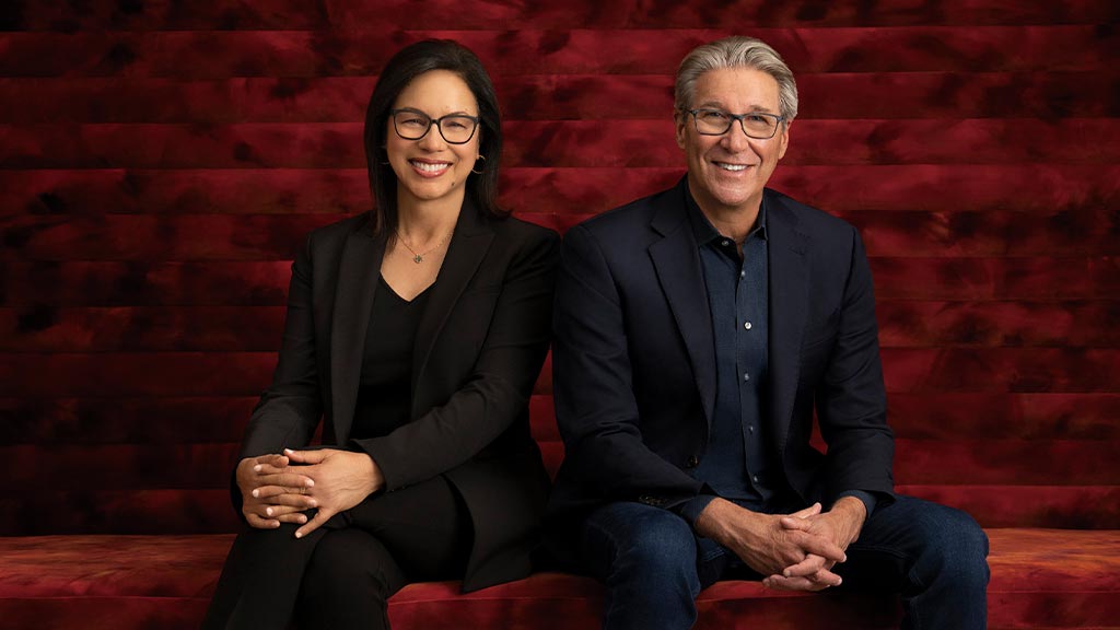 Diane Hoskins and Andy Cohen are sitting on a couch posing.