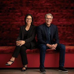 Diane Hoskins and Andy Cohen are sitting on a couch posing.